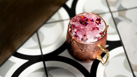 A copper Moscow mule mug filled with a pink cocktail and topped with dried rose petals, set on a black and white patterned tile surface.