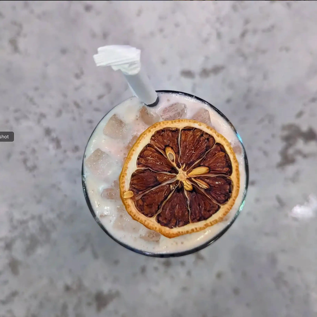 Overhead view of a dehydrated lemon wheel floating on a creamy iced cocktail with a white straw, on a grey concrete bar top