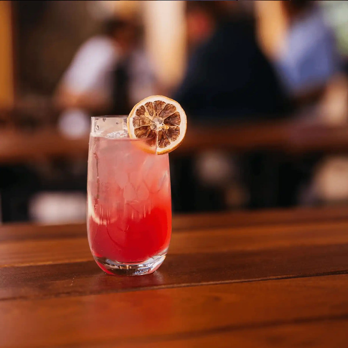 Dehydrated lemon wheel perched on the rim of a tall pink cocktail over ice, on a wooden bar top with guests blurred in the background