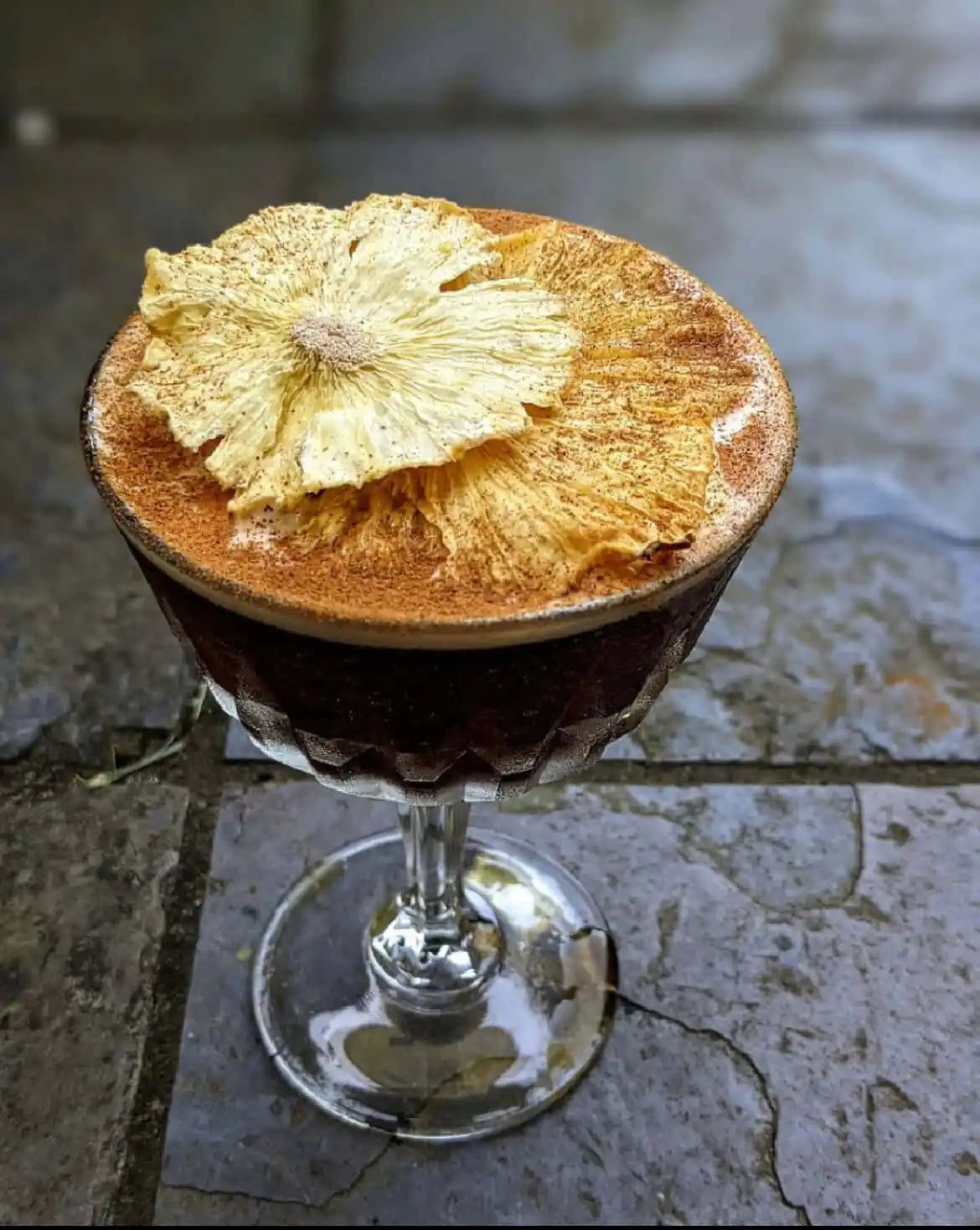Dehydrated pineapple flower resting on the crema of an espresso martini dusted with cinnamon, served in a faceted coupe on slate