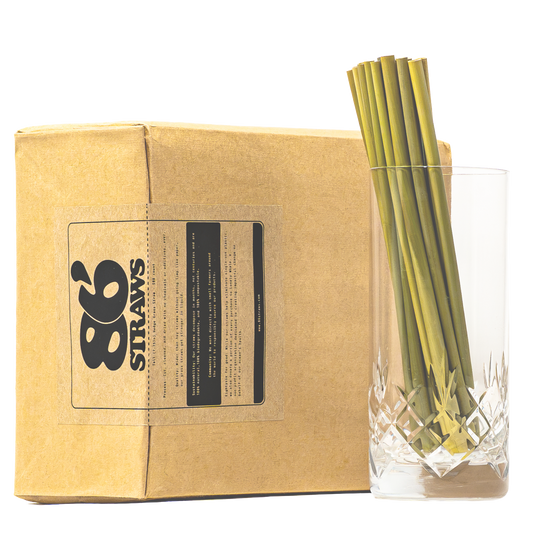 A kraft paper box with a black 86 Straws logo label sits next to a clear cut-glass tumbler holding several green straws, photographed on a white background.
