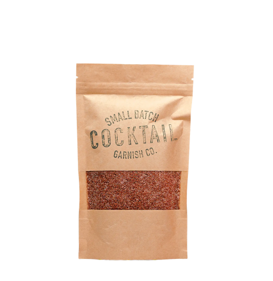 A small kraft paper stand-up pouch with a clear window showing reddish-brown strawberry sugar cocktail rimmer inside. The pouch has dark printed text reading 'SMALL BATCH COCKTAIL GARNISH CO.