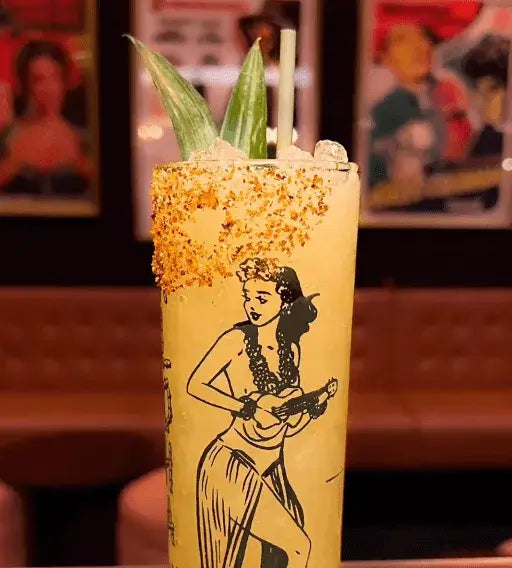 a tiki glass with an outer rim of blood orange and himalayan sea salt with two pineapple leaves and a hedge grass straw
