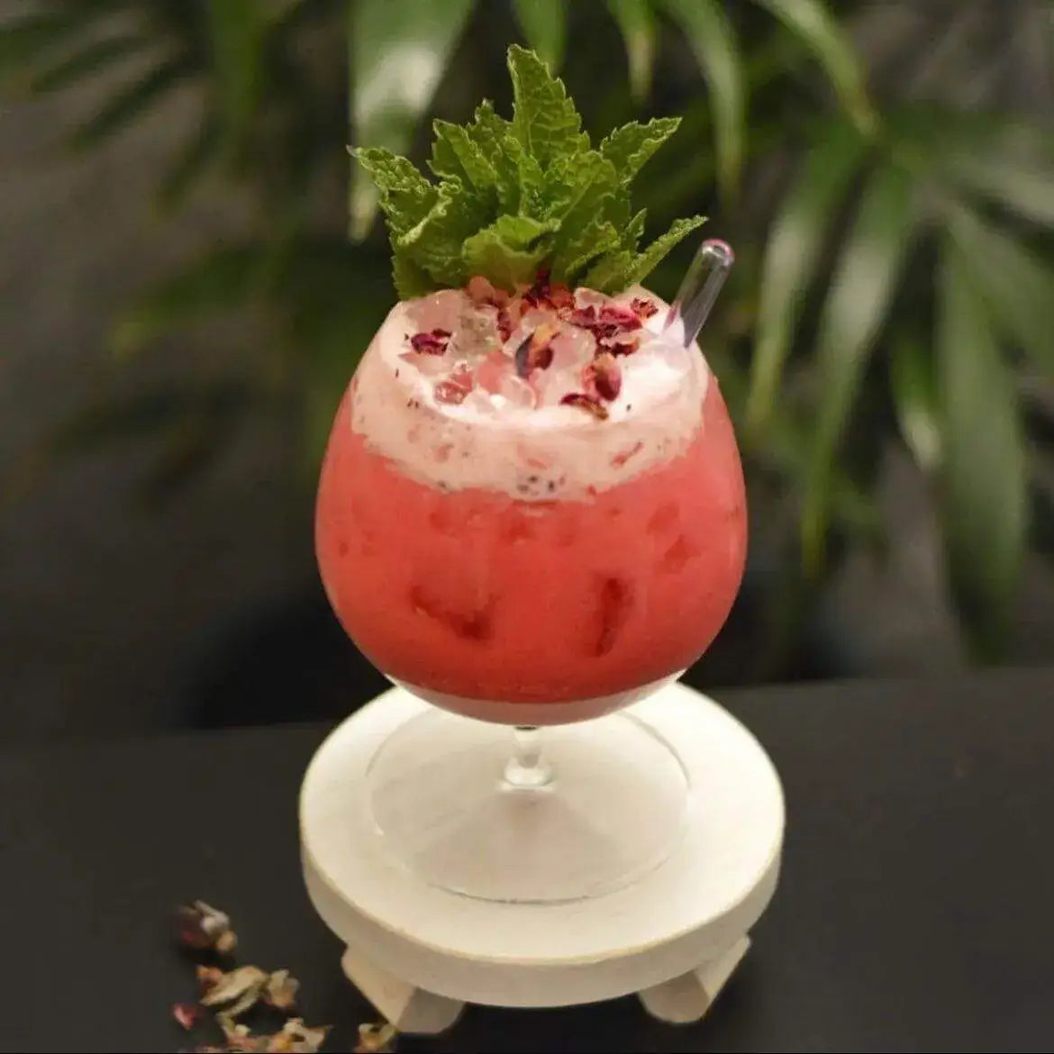 Crushed dried rose petals scattered over the foam of a red swizzle-style cocktail with a mint bouquet and glass straw, set on a white riser against dark foliage