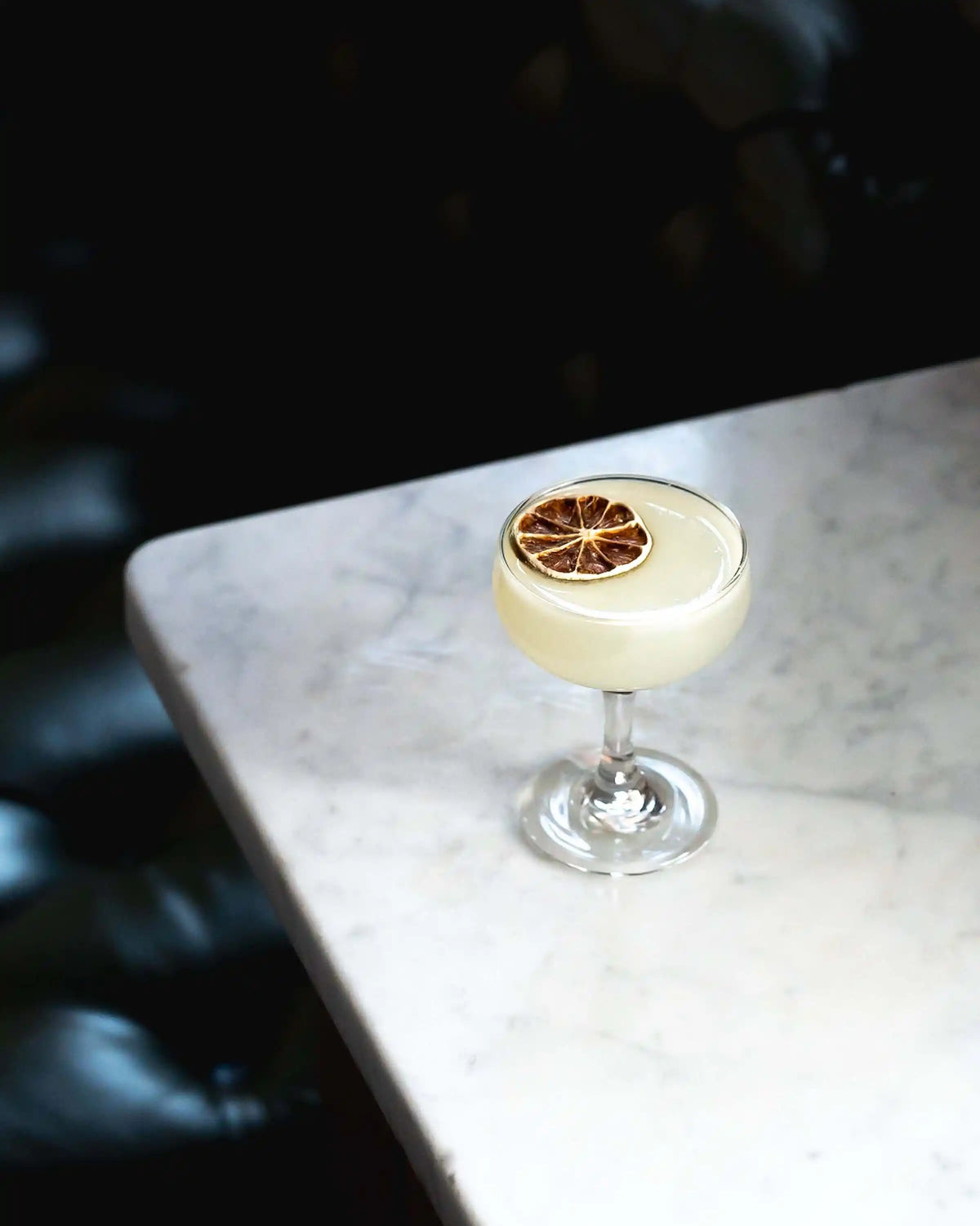 Dehydrated lime wheel floating on a pale yellow coupe cocktail at the corner of a white marble bar top in a dimly lit bar