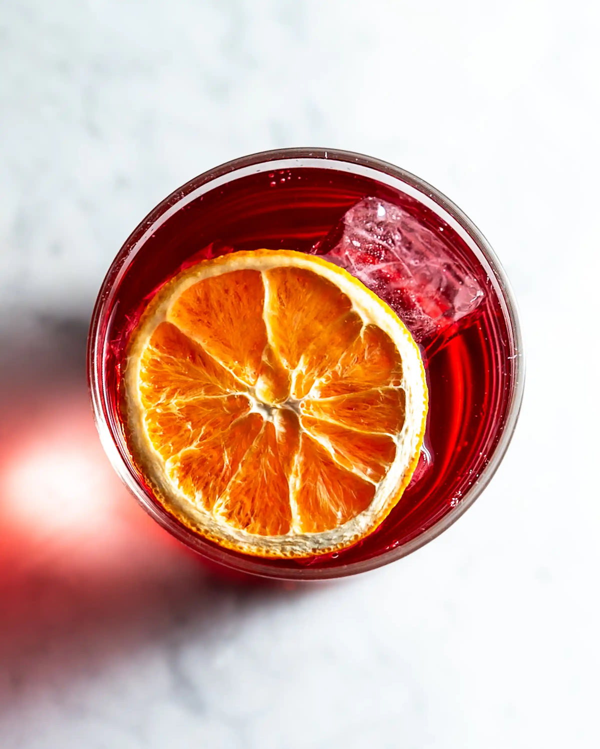 Overhead view of a dehydrated orange wheel floating on a deep red Negroni with a single large ice cube, on white marble