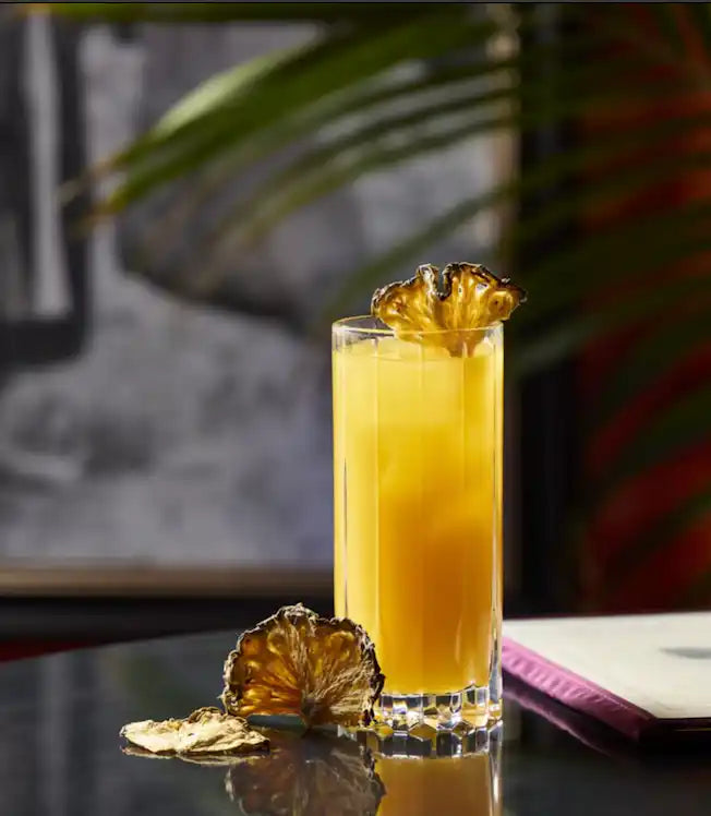 Dehydrated pineapple wheel garnishing a tall pineapple cocktail in a fluted highball glass, with two loose pineapple slices on a dark reflective bar table