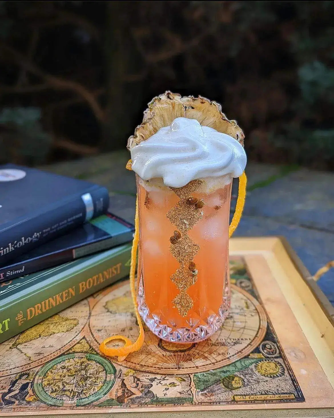 Dehydrated Chamomile syrup based beverage with dehydrated pineapple wheel garnish standing behind a swirl of whipped cream on a tall orange tiki cocktail, with a long orange peel twist and a stack of cocktail books behind