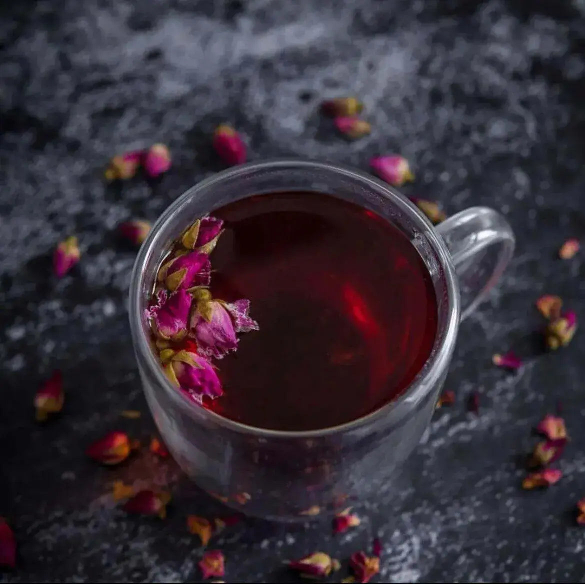 Whole dried rosebuds floating in a deep red rose tea in a clear glass mug, with loose pink rosebuds scattered on dark slate