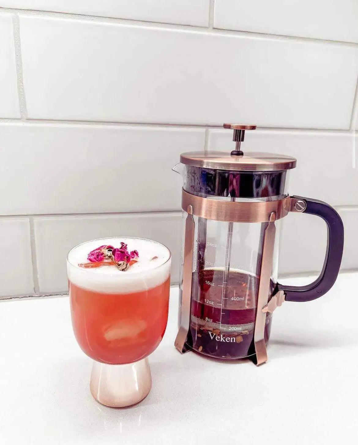 Foamed pink rose cocktail garnished with dried rosebuds beside a rose infusion in a French press